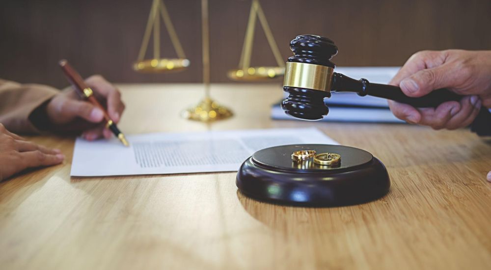 Gavel and wedding rings on table, legal documents being signed in divorce proceeding, justice scales in background.
