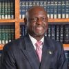 Smiling man in suit with pink tie stands in front of bookshelf filled with legal books.