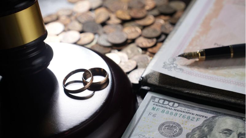 Wedding rings, gavel, currency, and documents symbolize divorce and financial settlement.