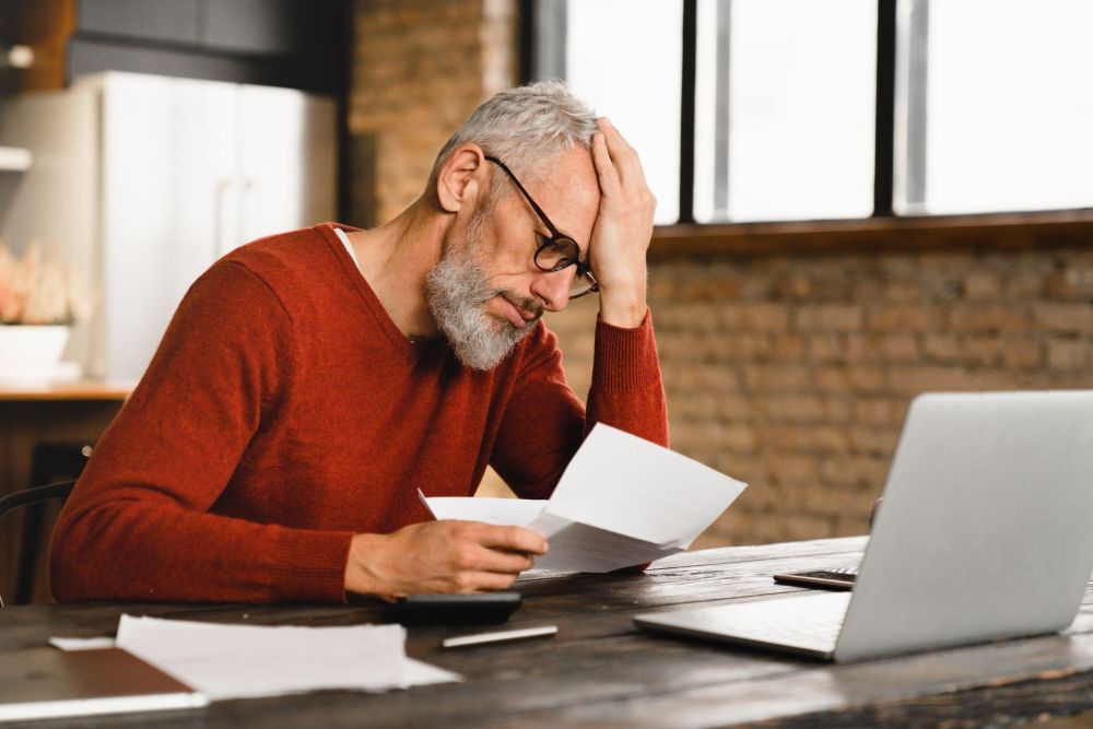 Man in red sweater looks stressed while reviewing documents at a laptop in a modern office.