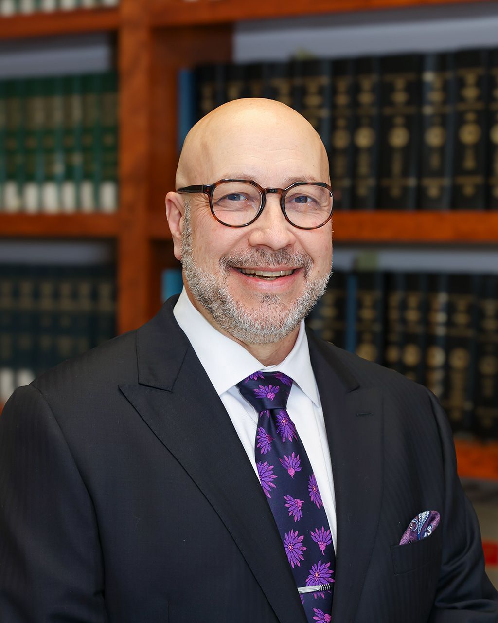 Christopher Dunn in suit with glasses and purple tie smiling in a library with shelves of books.