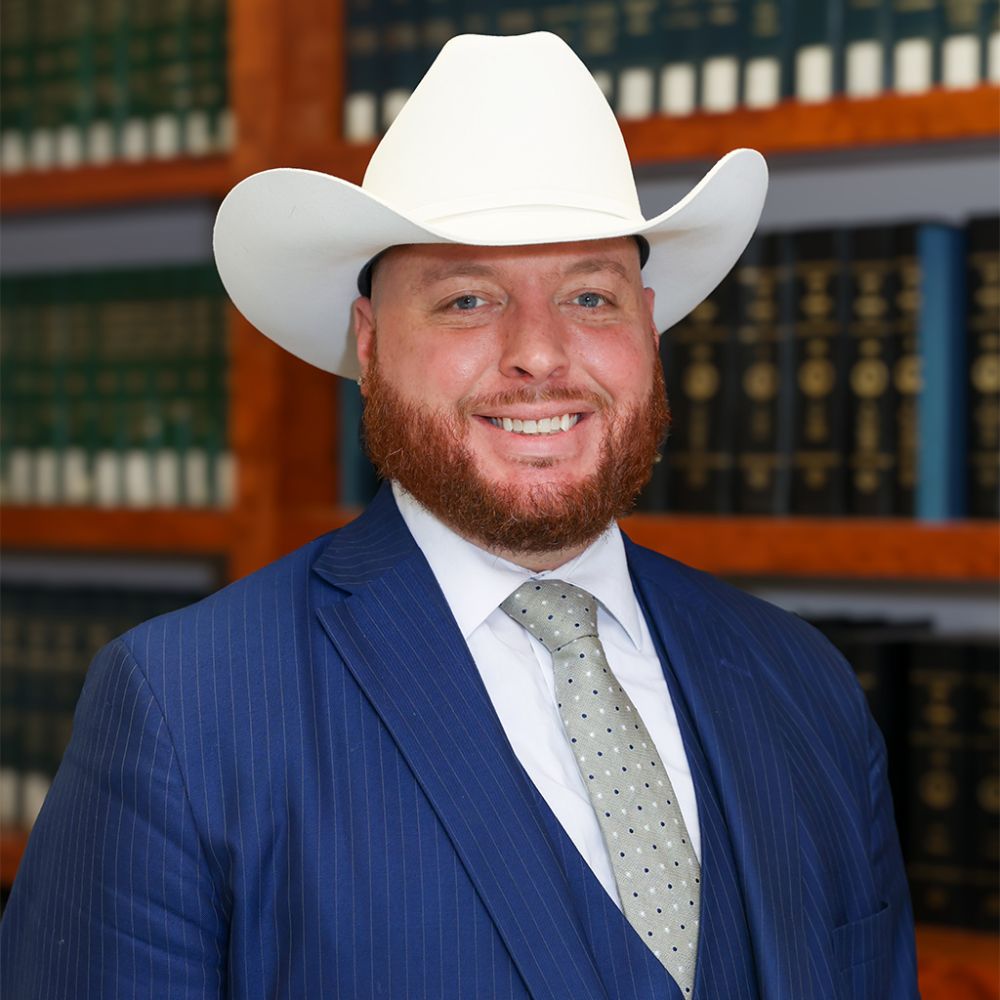 Christopher Daniels in white cowboy hat and blue suit smiling, standing in front of bookshelves filled with law books.