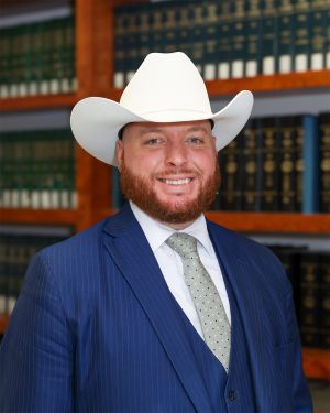 Christopher Daniels in white cowboy hat and blue suit smiling, standing in front of bookshelves filled with law books.