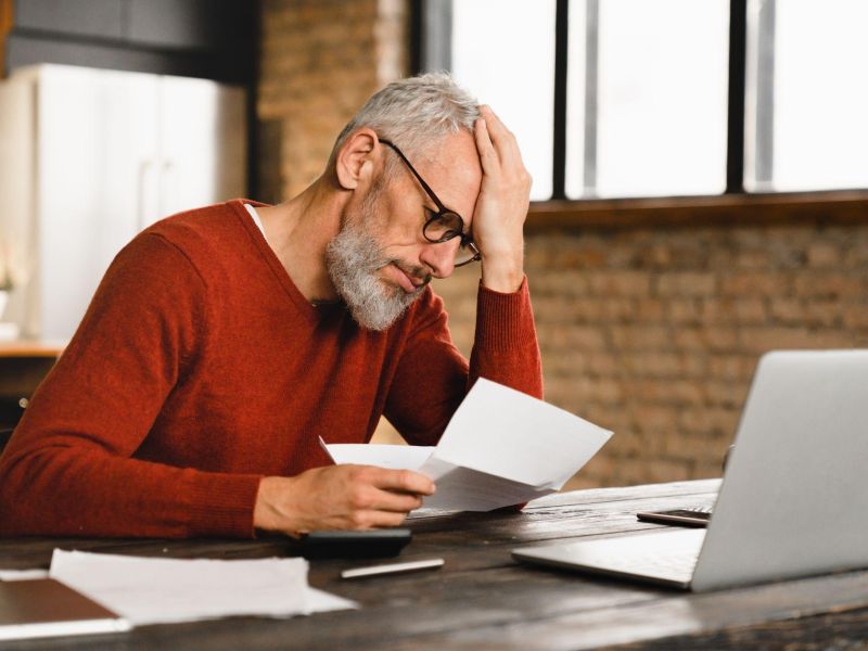 Man in red sweater looks stressed while reviewing documents at a laptop in a modern office.