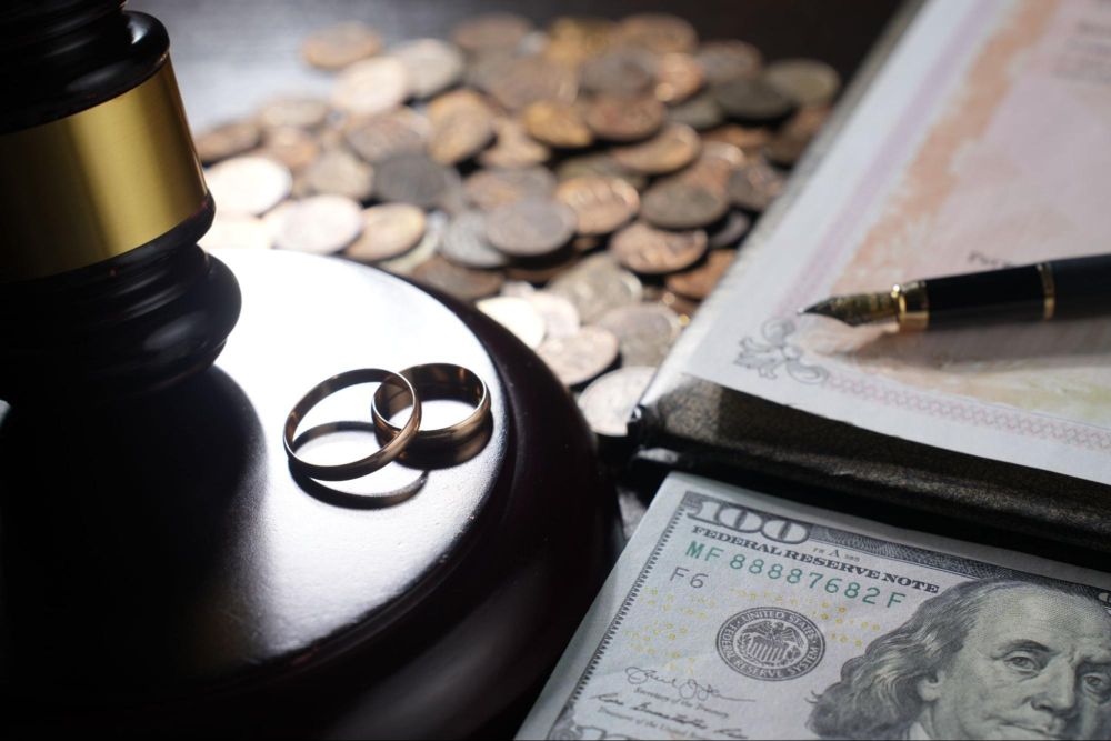 Wedding rings, gavel, currency, and documents symbolize divorce and financial settlement.