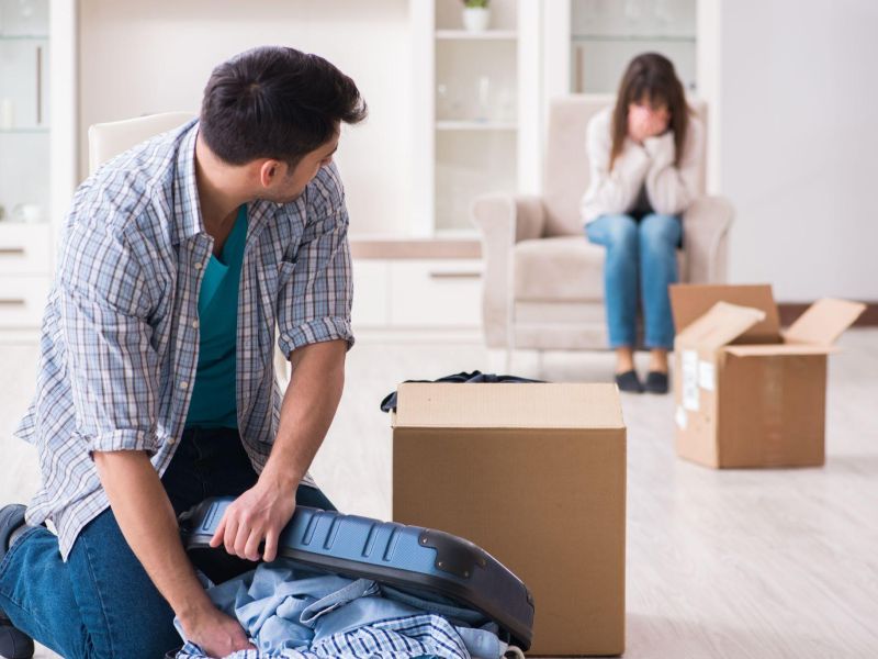Man packing suitcase, while woman sits upset in background, surrounded by moving boxes in a room.