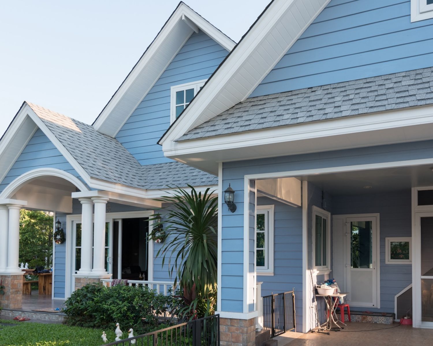 Blue suburban house with white trim, featuring a front porch, green lawn, and columns under a clear sky.