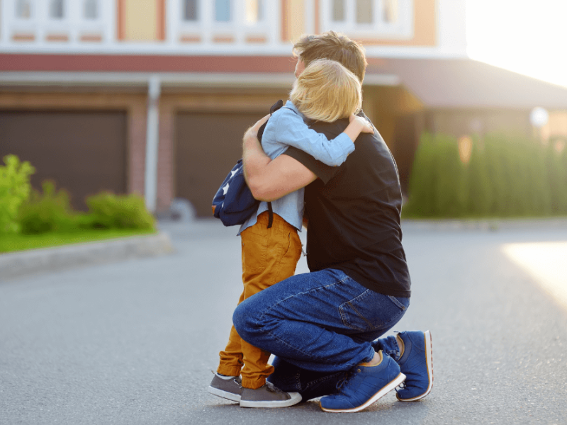A child hugs an adult kneeling in a driveway, displaying affection. The setting includes a suburban background with garages and greenery, illuminated by warm sunlight.
