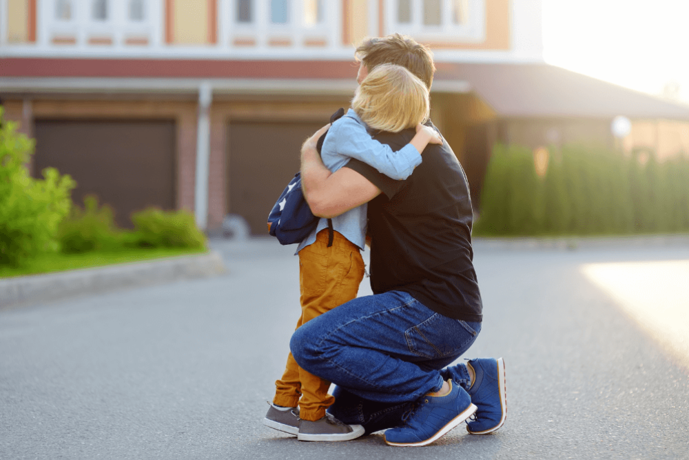 A child hugs an adult kneeling in a driveway, displaying affection. The setting includes a suburban background with garages and greenery, illuminated by warm sunlight.