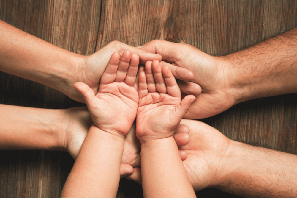 Several hands of different sizes, including a child's, are stacked together in a gesture of unity, resting on a textured wooden surface, symbolizing connection and support.