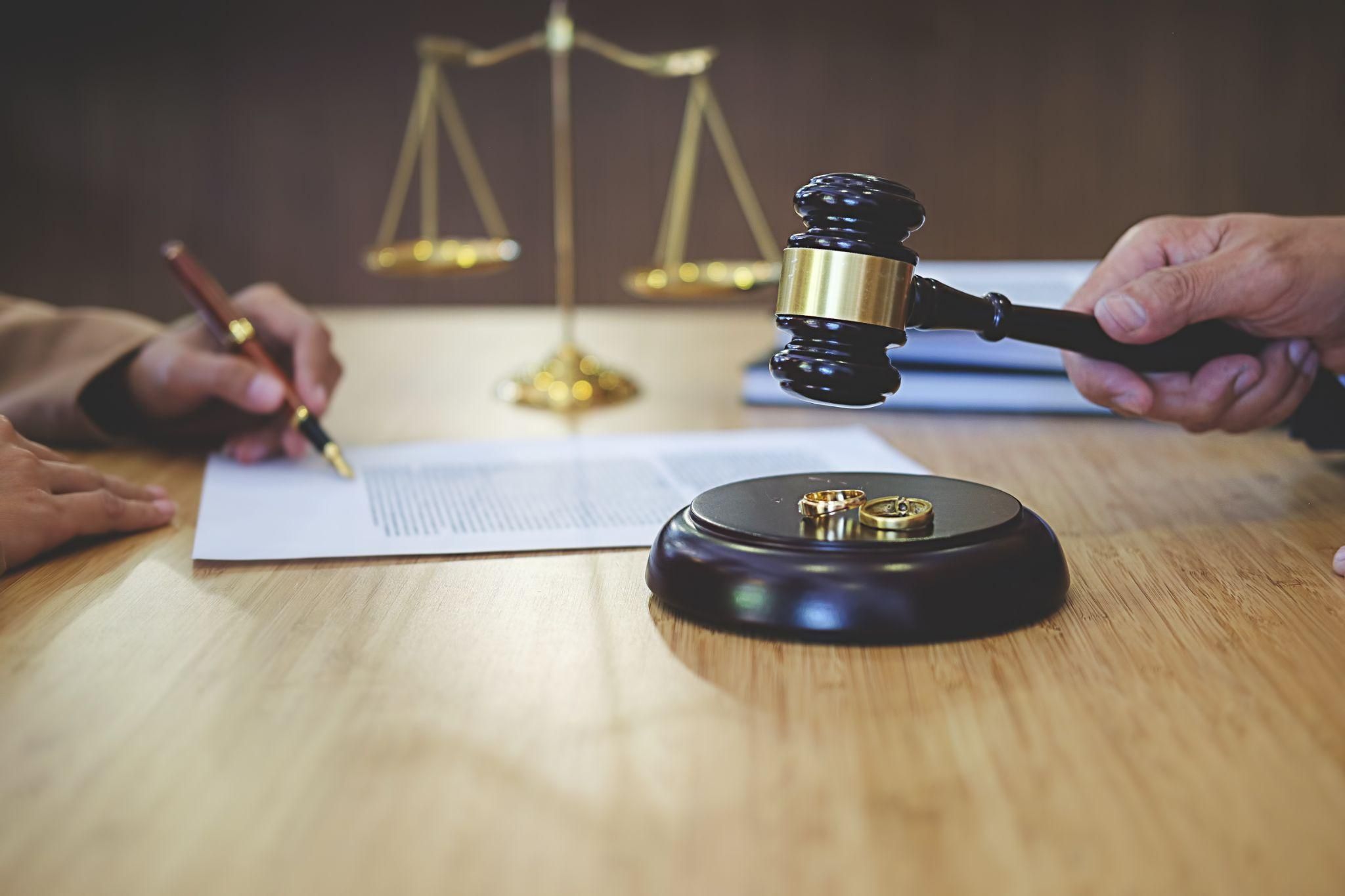 Gavel and wedding rings on table, legal documents being signed in divorce proceeding, justice scales in background.