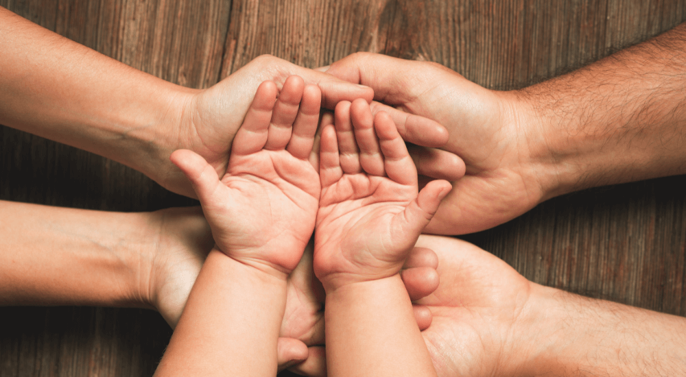 Several hands of different sizes, including a child's, are stacked together in a gesture of unity, resting on a textured wooden surface, symbolizing connection and support.