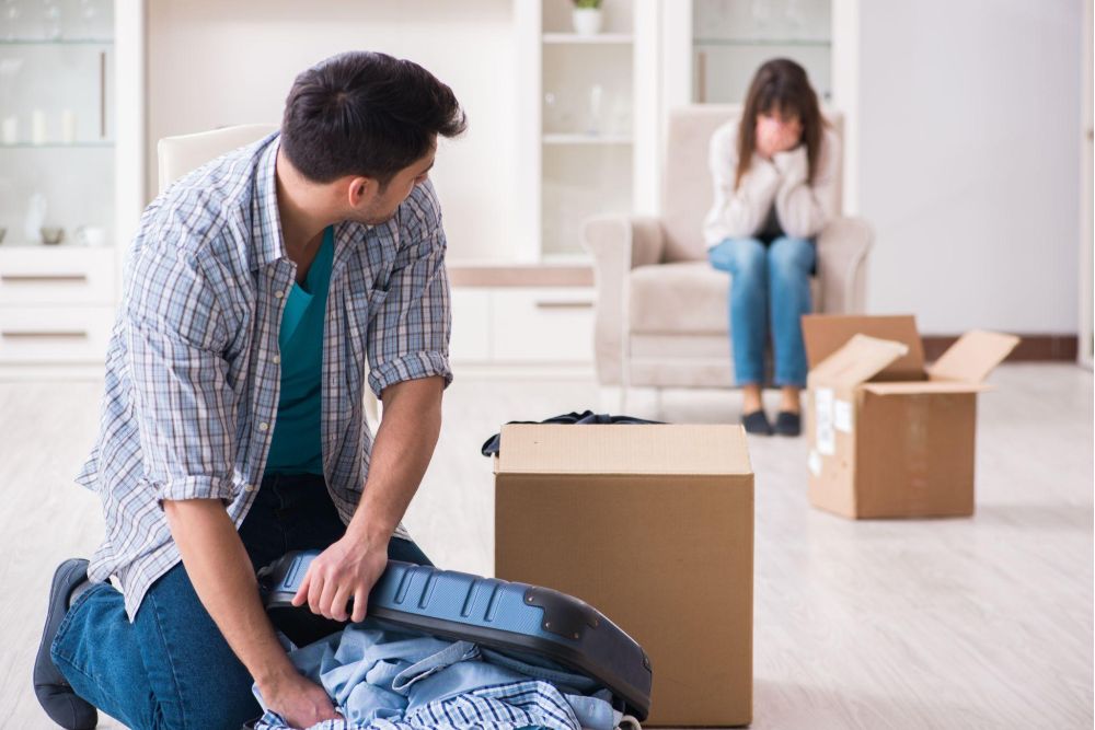 Man packing suitcase, while woman sits upset in background, surrounded by moving boxes in a room.