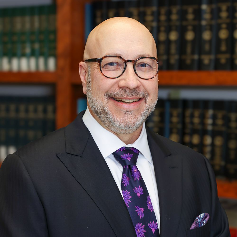 Christopher Dunn in suit with glasses and purple tie smiling in a library with shelves of books.