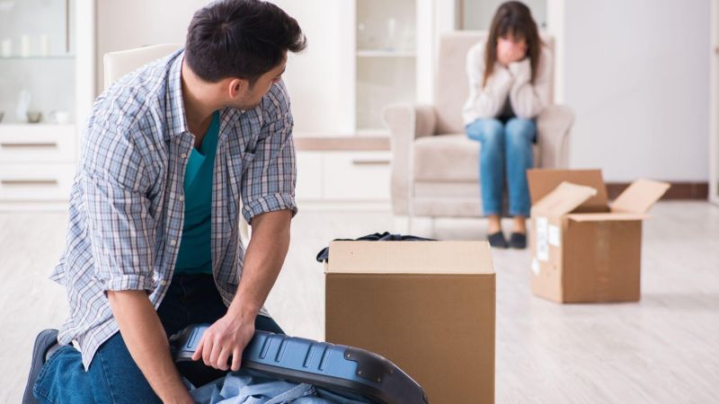 Man packing suitcase, while woman sits upset in background, surrounded by moving boxes in a room.