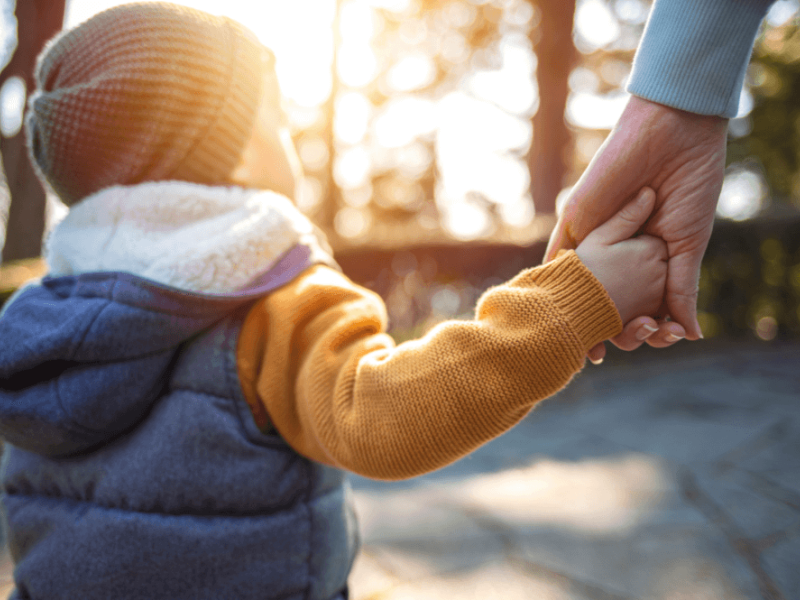 A child in a colorful sweater and hat holds hands with an adult. They are walking through a sunny, outdoor setting with soft light filtering through trees, suggesting warmth and connection.