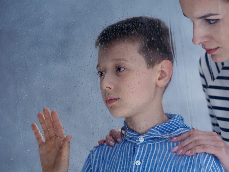 Boy and woman looking through rainy window, expressing contemplation and connection.