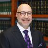 Christopher Dunn in suit with glasses and purple tie smiling in a library with shelves of books.