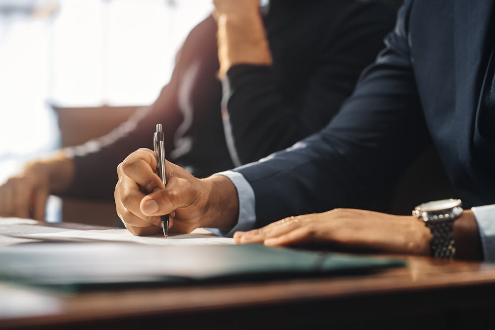 man sitting at desk signing papers with suit on and watch on his wrist 