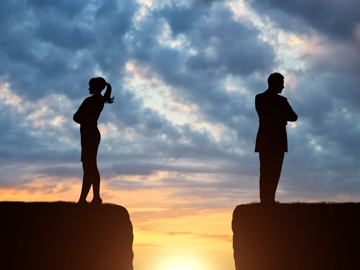 Silhouetted couple stands on opposite cliffs at sunset, symbolizing separation and conflict under a cloudy sky.