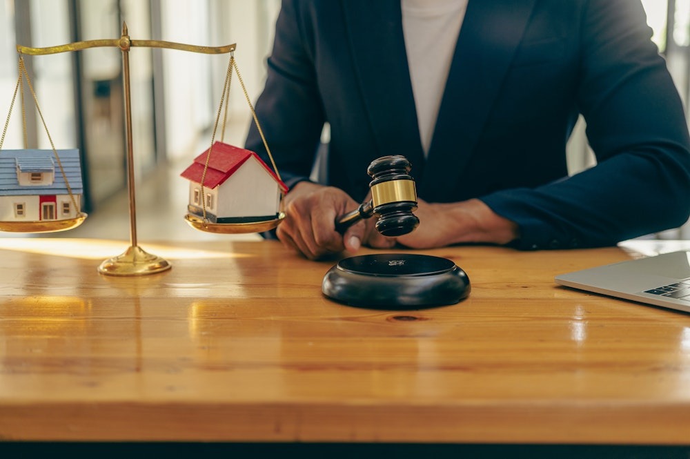 Person weighing house models on a scale, with a gavel on the desk symbolizing real estate decision making.