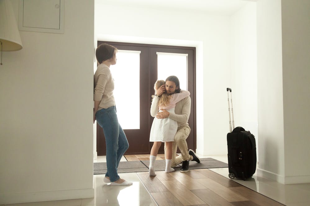 A man kneels to embrace a young girl, showing affection. A woman leans against a wall, while a suitcase stands nearby in a bright, modern entryway.