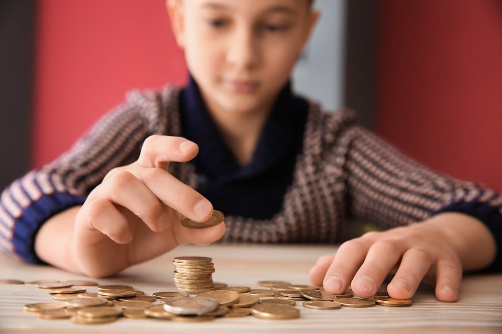 A child stacks coins on a table with focused concentration. Surrounding them are more scattered coins, and the background features a softly blurred red wall.
