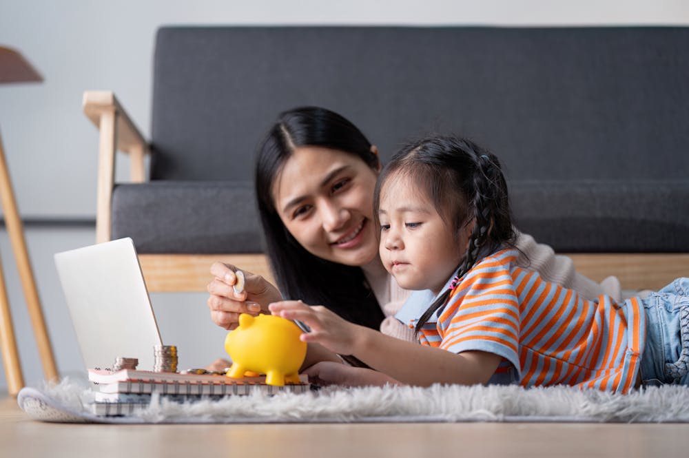 A mother and child are lying on a soft rug, engaging with a laptop. The mother helps the child with a yellow piggy bank, surrounded by coins, in a cozy living room.