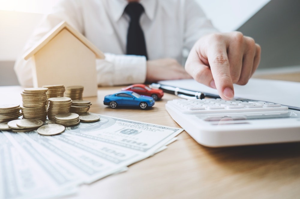 Person calculating financial assets with coins, model cars, and house on desk. Budget planning concept.