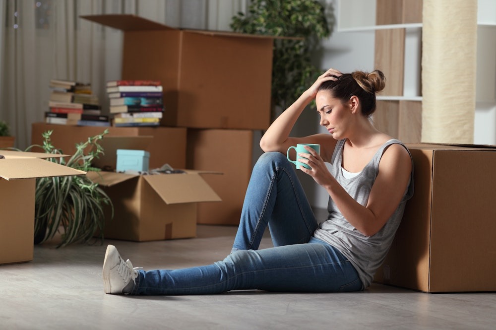 Woman sitting on floor with coffee among moving boxes, appearing thoughtful and tired in a new home setup.