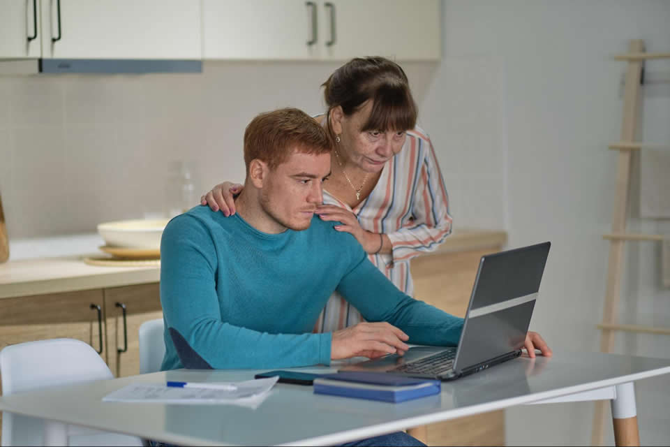 Woman guiding man working on a laptop in a modern kitchen setting.