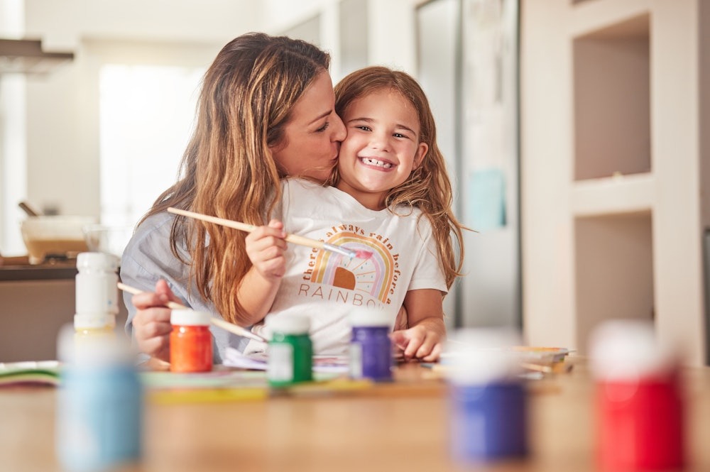 Mother and daughter enjoying painting together, smiling warmly in a home setting with colorful paints on the table.