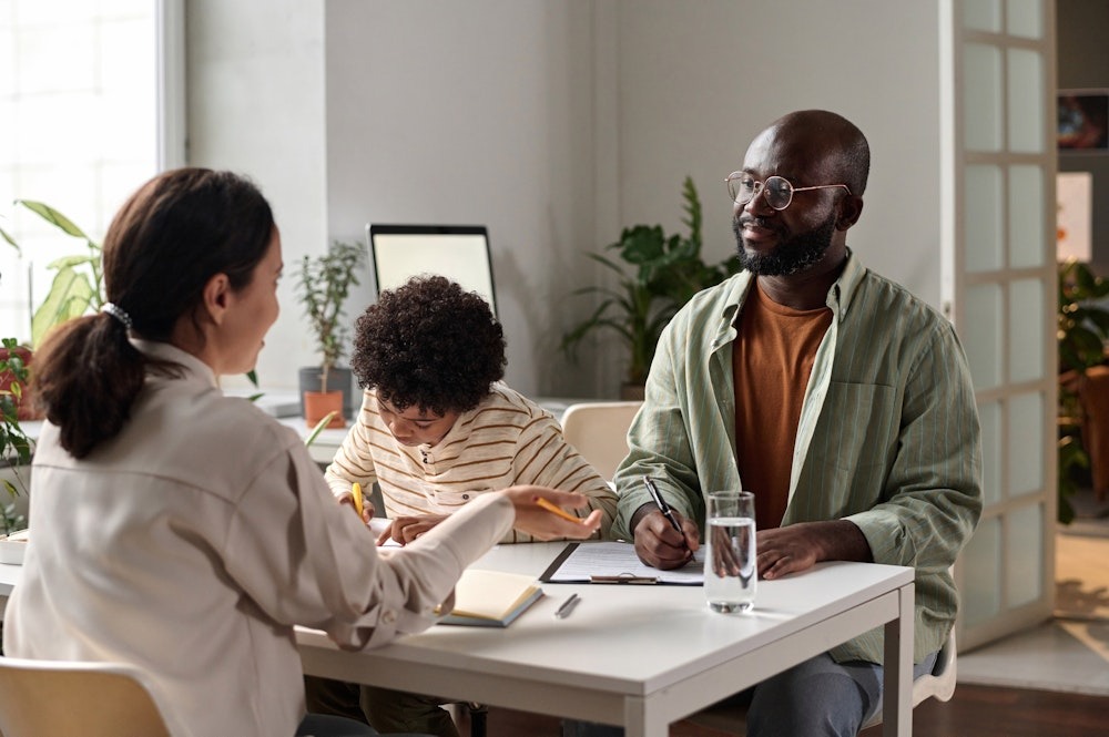 Family working together at a table with papers, glass of water, and plants in a bright room.