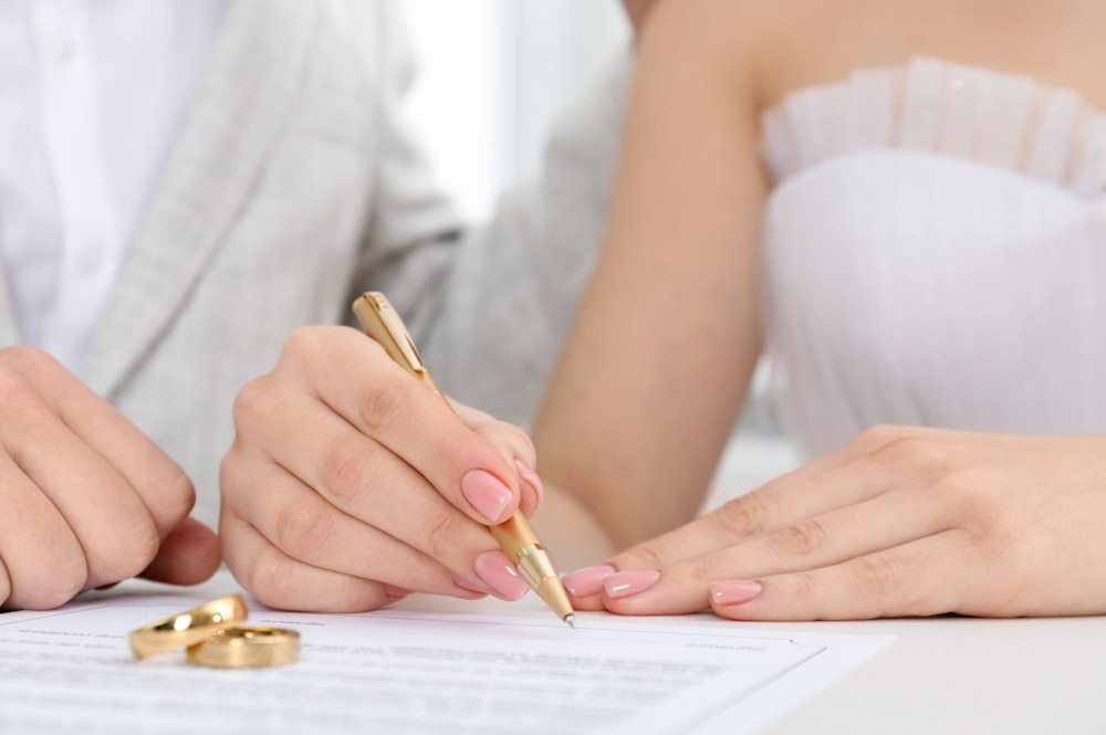 Bride signing wedding certificate with gold pen, close-up of hands and rings on the table.