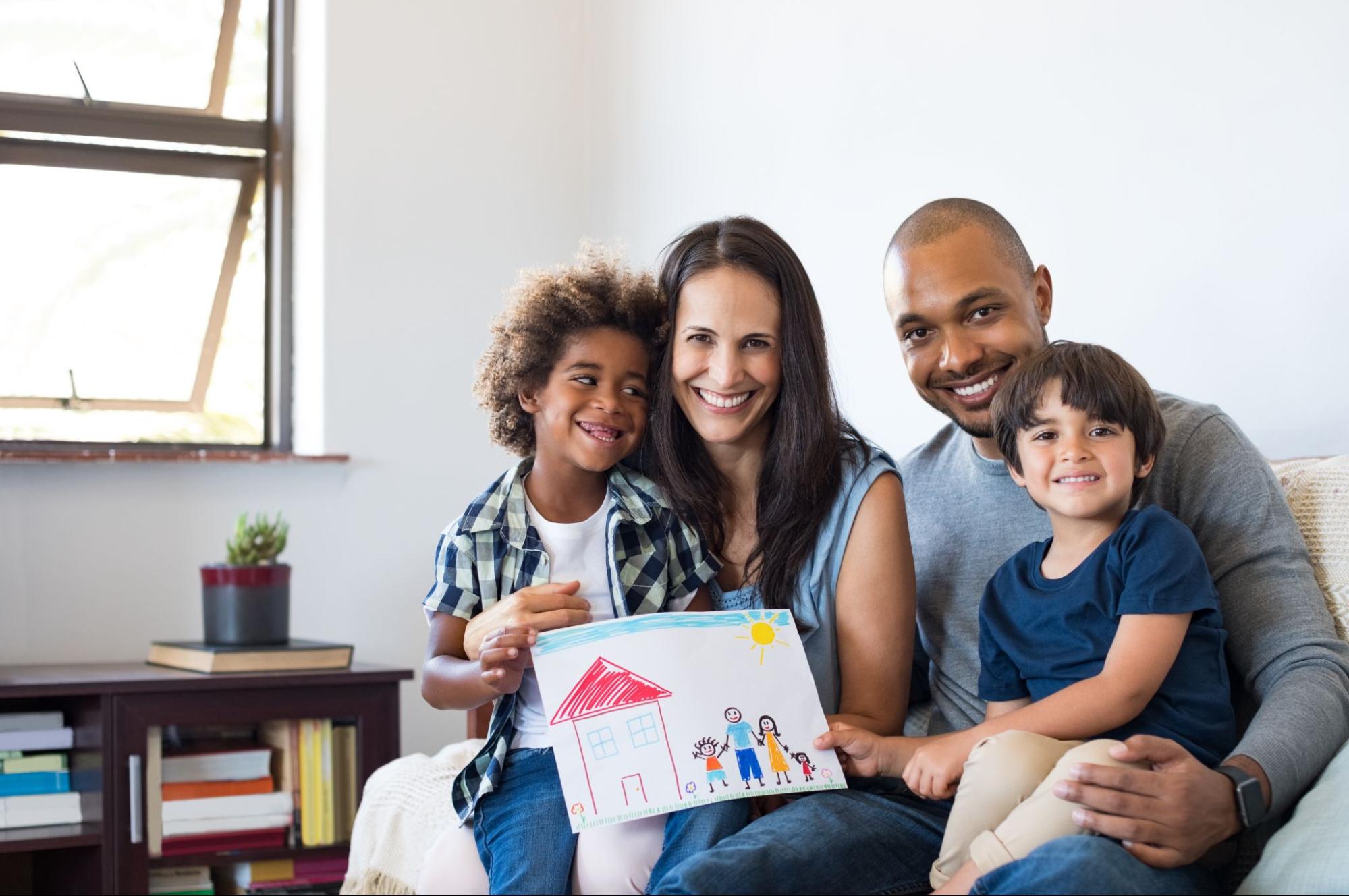 Happy family of four sitting together, smiling and holding a child's drawing of a house and family in a cozy living room.