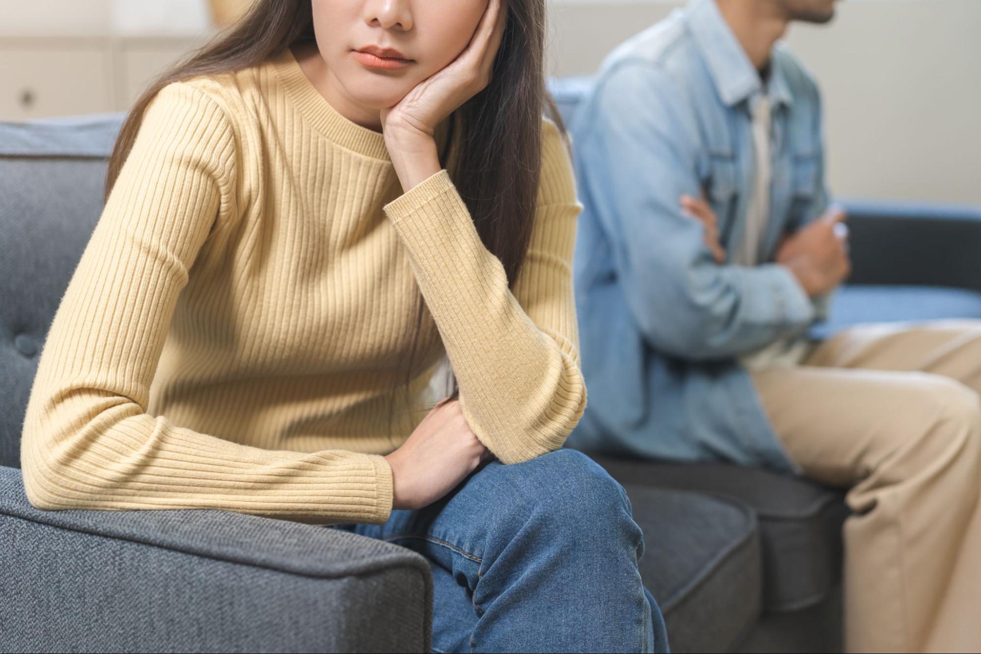 Couple sitting apart on couch after disagreement, woman in yellow sweater looks upset while man crosses arms.