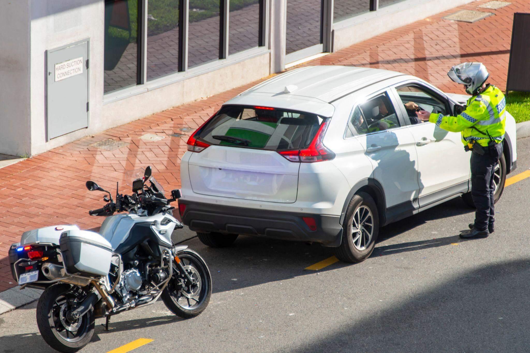 a police officer standing next to a parked car, his motorcycle is behind the car in a sunny day